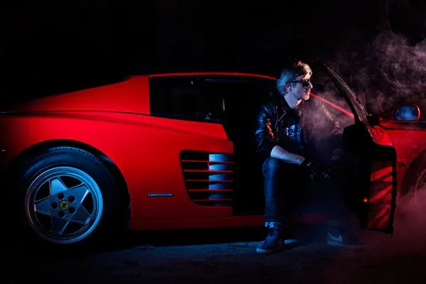 Kavinsky assis à la portière d'une Ferrari Testarossa rouge dans une lumière rouge et bleue