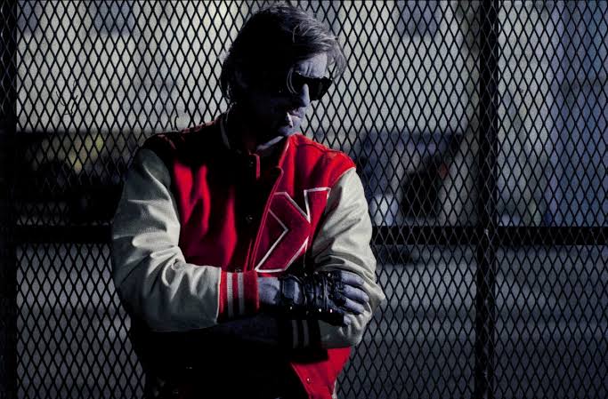 Kavinsky en veste teddy rouge et blanche au logo K, lunettes noires, adossé à un grillage