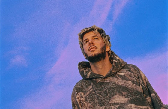 Fakear vu en contre-plongée, sweat camouflage, sur un ciel bleu et rose Titre : Fakear – portrait presse 2026